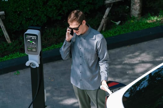 Young man travel with EV electric car charging in green sustainable city outdoor garden in summer shows urban sustainability lifestyle by green clean rechargeable energy of electric vehicle innards