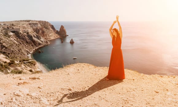 Side view a Young beautiful sensual woman in a red long dress posing on a rock high above the sea during sunrise. Girl on the nature on blue sky background. Fashion photo.