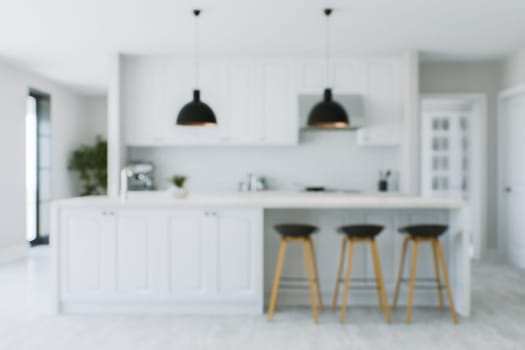 The kitchen background is out of focus. Blurred white kitchen with island, lamps, bar stools and kitchen appliances. 3D rendering
