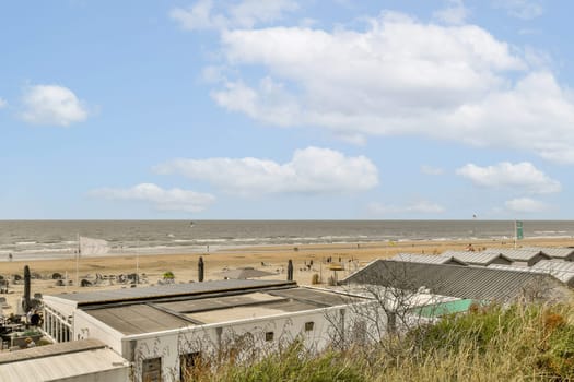 a beach with some buildings in the fore, and people walking on the other side of the beach looking out at the ocean