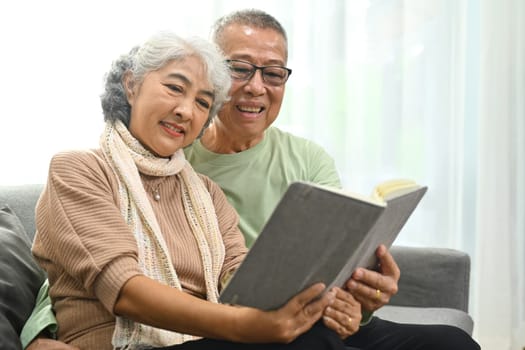 Beautiful senior couple embracing and reading book on sofa while spending time together at home.