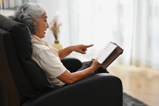 Carefree senior woman sitting on the armchair and reading book. Elderly people lifestyle and relaxation concept.