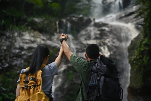 Rear view of young couple travelers with backpacks standing with raised hands in front of waterfall.