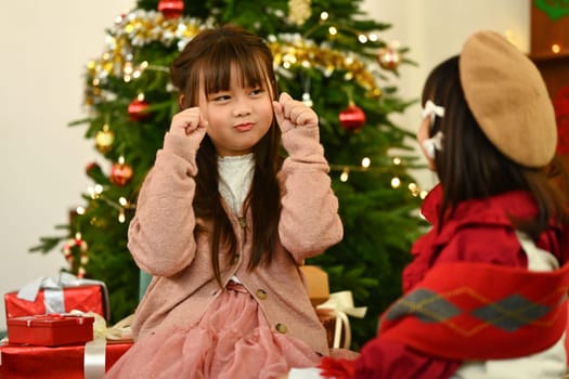 Cheerful little girls in warm winter clothes having fun and playing together near decorated Christmas tree.