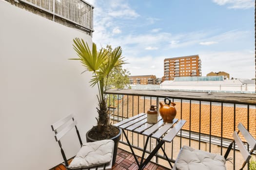 a balcony with two chairs and an orange vase on the table, in front of a white wall behind it