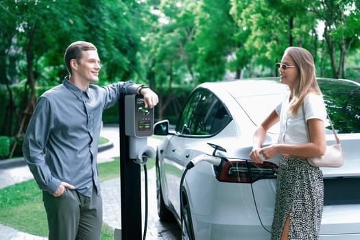 Young couple travel with EV electric car charging in green sustainable city outdoor garden in summer shows urban sustainability lifestyle by green clean rechargeable energy of electric vehicle innards