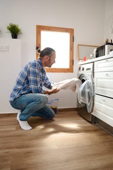 Young adult handsome Caucasian man doing the laundry at home, taking off laundered clothing out of a washing machine. Washing clothes. The concept of washing and cleanliness. Household and chores