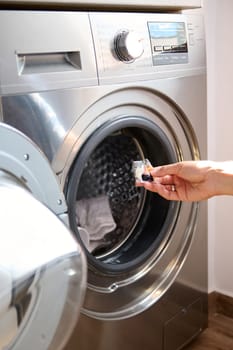 Close-up housewife hand putting a laundry detergent or washing pod into a modern automatic washing machine. Hand holding eco gel in capsule. Washing clothes. The concept of washing and cleanliness