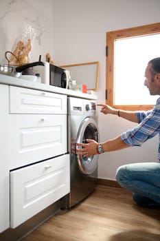 Close-up of a young adult handsome Caucasian man loading dirty clothes, adding laundry detergent, and turning on the automatic washing machine. Washing and cleanliness concept. Housekeeping and chores