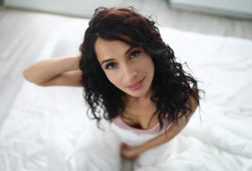 Portrait of brunette woman with a slight smile on background of white bed. Morning awakening and positive mood concept