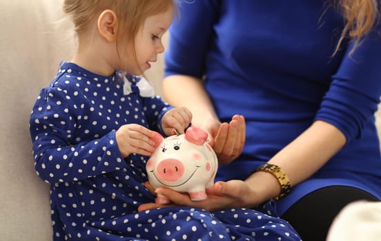 Child little girl arm putting pin money coins into happy pink faced piglet slot closeup. Making effective future needs savings collect dollar gift benefit present home leisure concept