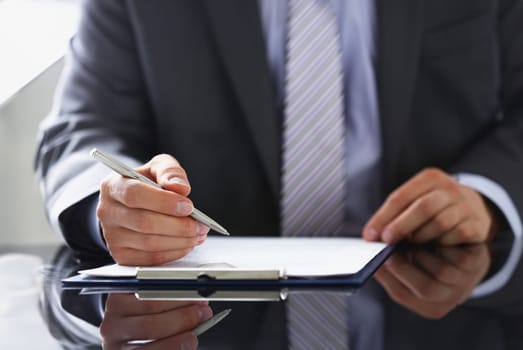 Male arm in suit and tie fill form clipped to pad with silver pen closeup. Sign gesture read pact sale agent bank job make note loan credit mortgage investment finance chief legal law concept