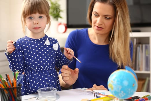 Blond smiling little girl hold in arm brush drawing something together with mom. Beautiful female young artist, minor baby-sitter, art joy, juvenile development, parent lifestyle, youth, painter