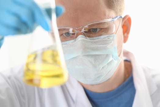 A portrait of a young surgeon chemist doctor looks at a container with a yellow liquid. Mask is fought with viruses and a vaccine for vaccines against diseases.