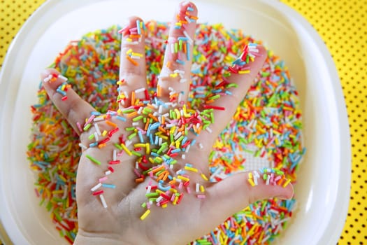 Children hand full of little colorful sweets, plate over yellow