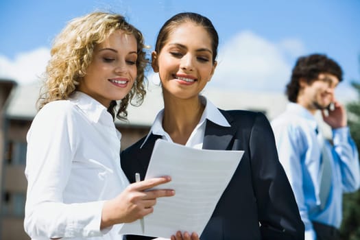 Cute smiling female students reading the documents outside and handsome young man talking by the phone on the background