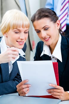 Portrait of two confident business ladies sitting at the table and discussing business documents looking into them with closed laptop near by
