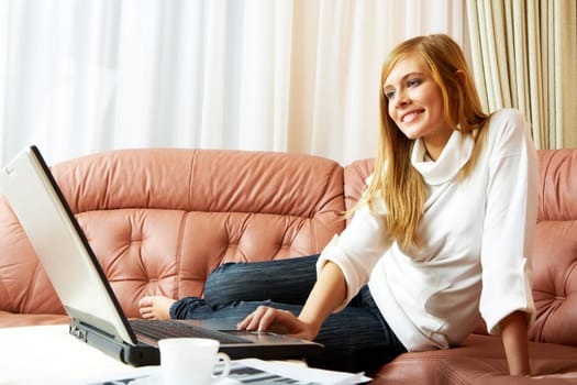 Image of pretty girl in home clothes looking at laptop monitor while sitting on leather sofa