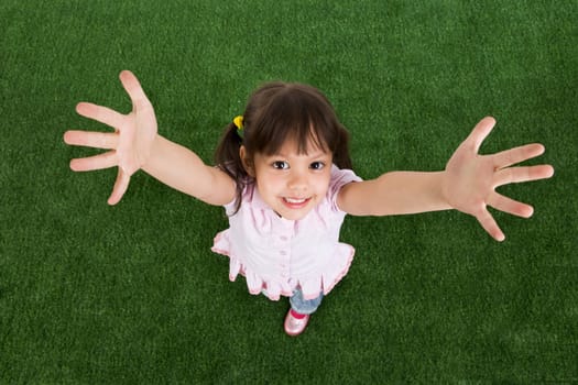 Above view of happy child standing on green grass and raising her arms to camera