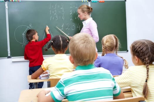 Photo of elementary student by blackboard looking at his teacher helping him