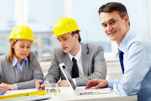 A manager working with laptop, looking at camera and smiling against his colleagues architects