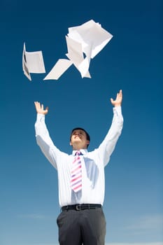 Portrait of successful businessman standing and tossing up papers on background of blue sky