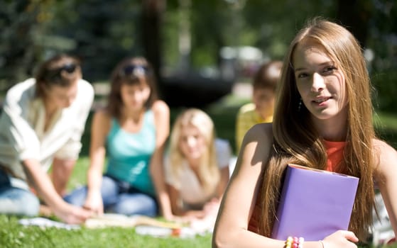 Portrait of cheerful girl with book in hands looking at camera on background of her friends