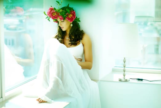 Beautiful woman in white dress sitting on window sill and looking through glass