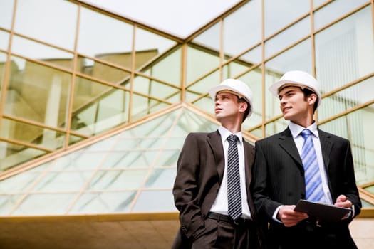 Photo of young engineers looking aside with modern building at background