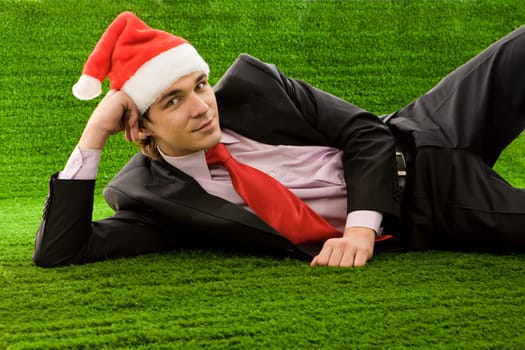 Image of satisfied businessman in santa cap lying on green grass and looking at camera