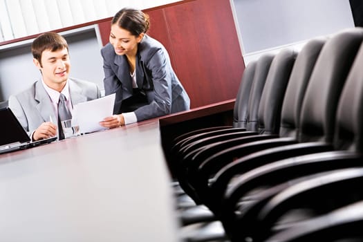 Image of a business man sitting and reading a document in the office with a smiling business lady standing near by