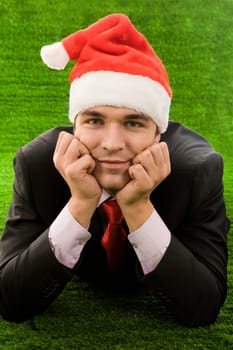 Businessman in santa cap touching his face by hands while relaxing on green grass