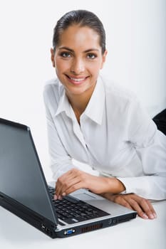 Isolated on white portrait of smiling suntanned woman sitting at the table touching keyboard of opened black laptop gazing at the camera