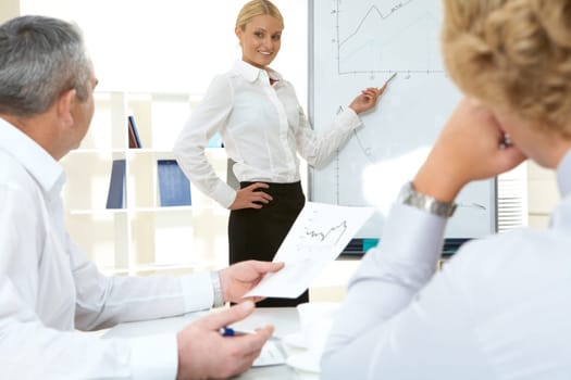 Photo of successful employee standing by whiteboard while her colleagues listening to her