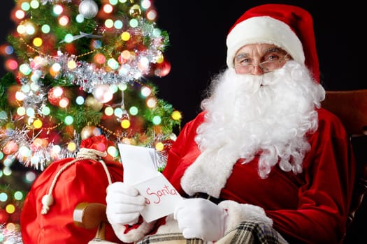Santa sitting at the Christmas tree, holding Christmas letters and looking at camera