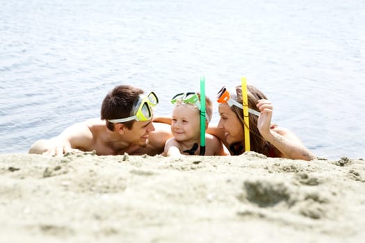 Portrait of cheerful couple and their son in diving equipment lying on sandy shore