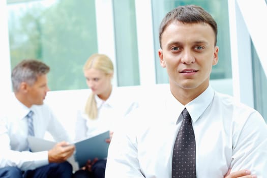 Confident businessman looking at camera at background of his communicating colleagues