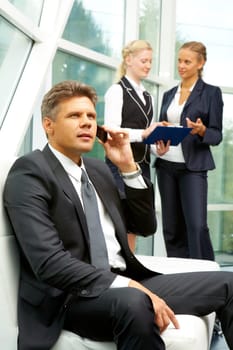 Confident businessman talking by mobile phone at background of his communicating colleagues