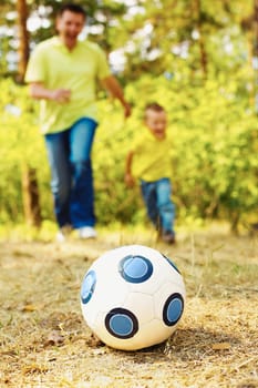 Image of ball on ground in park with running father and son at background