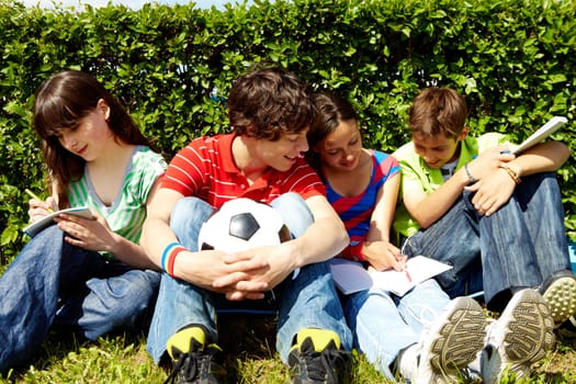 Portrait of modern teens in casual clothes sitting on green grass and writing in copybooks