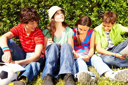 Portrait of inspired teens sitting on green grass and writing in copybooks