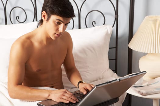 Young man sitting on bed and typing on laptop