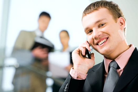 Portrait of a young handsome businessman speaking on the phone and looking at camera with smile in working environment