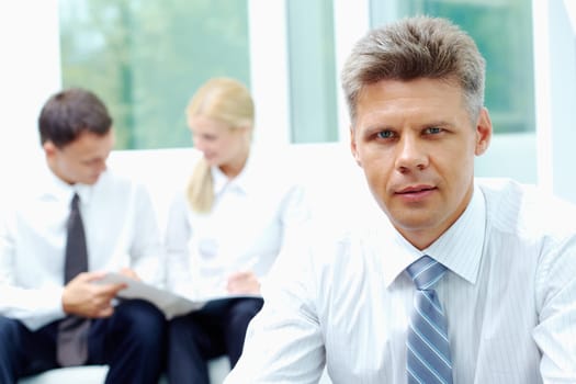 Confident businessman looking at camera at background of his communicating colleagues