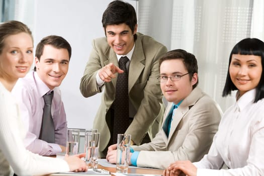 Image of friendly workteam sitting around table with their leader pointing at camera