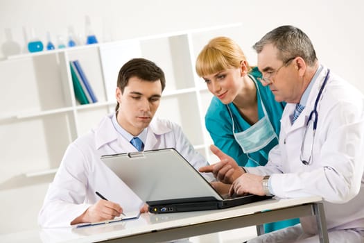 Photo of aged physician pointing at laptop display with two colleagues near by looking at it