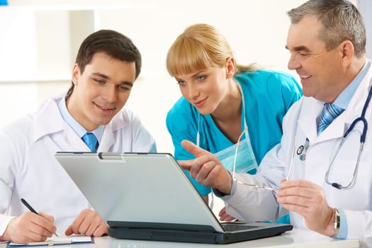 Photo of aged physician pointing at laptop display while explaining something to his colleagues