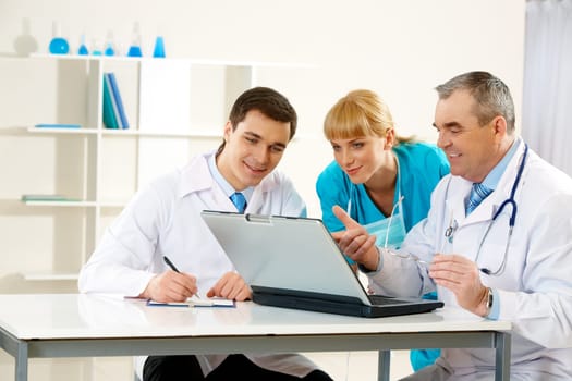 Photo of aged physician pointing at laptop display with two colleagues near by looking at it