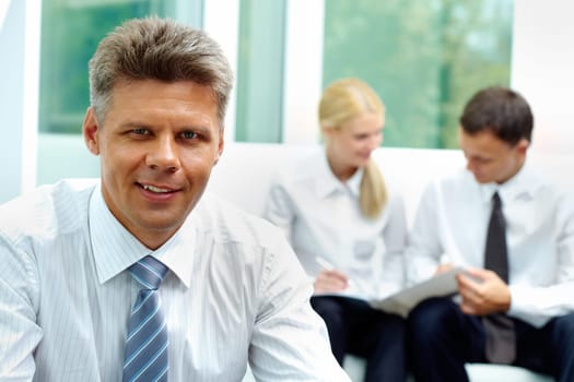 Confident businessman looking at camera at background of his communicating colleagues
