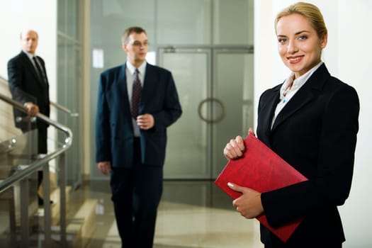 Smart beautiful businesswoman with red case in her hands and two men behind her going downstairs